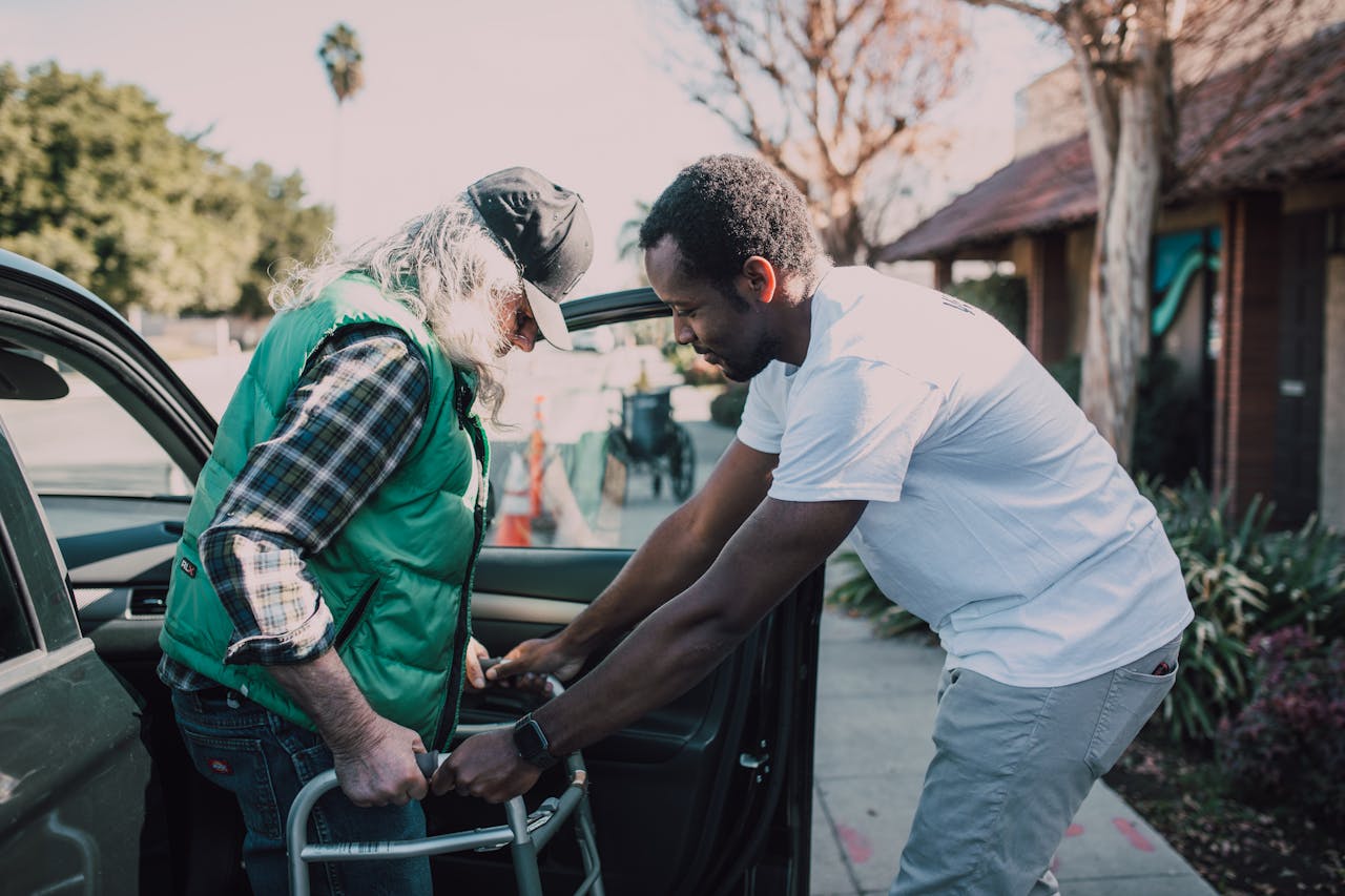 services-04 Young volunteer helping senior man with walker near a parked car, symbolizing care and support.